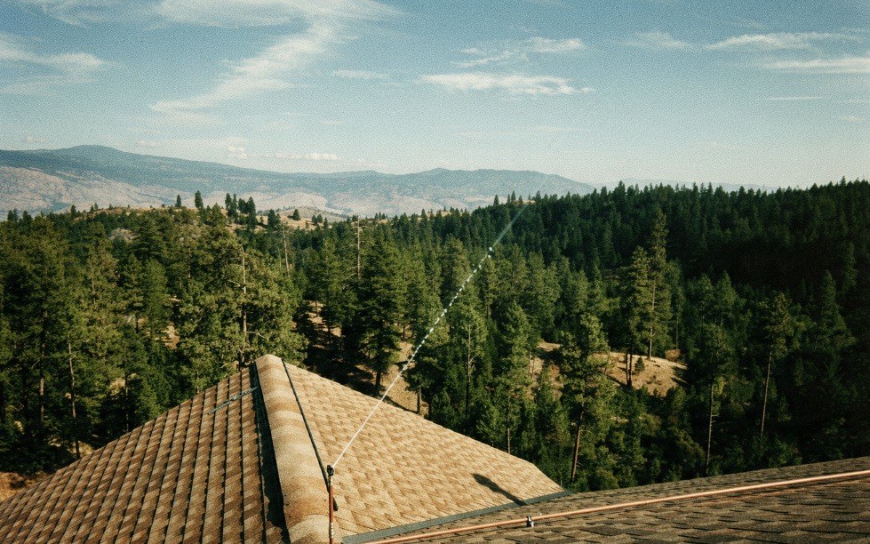 Rooftop wildfire sprinkler spraying an arc of water over a shingled roof with forested Okanagan hills behind
