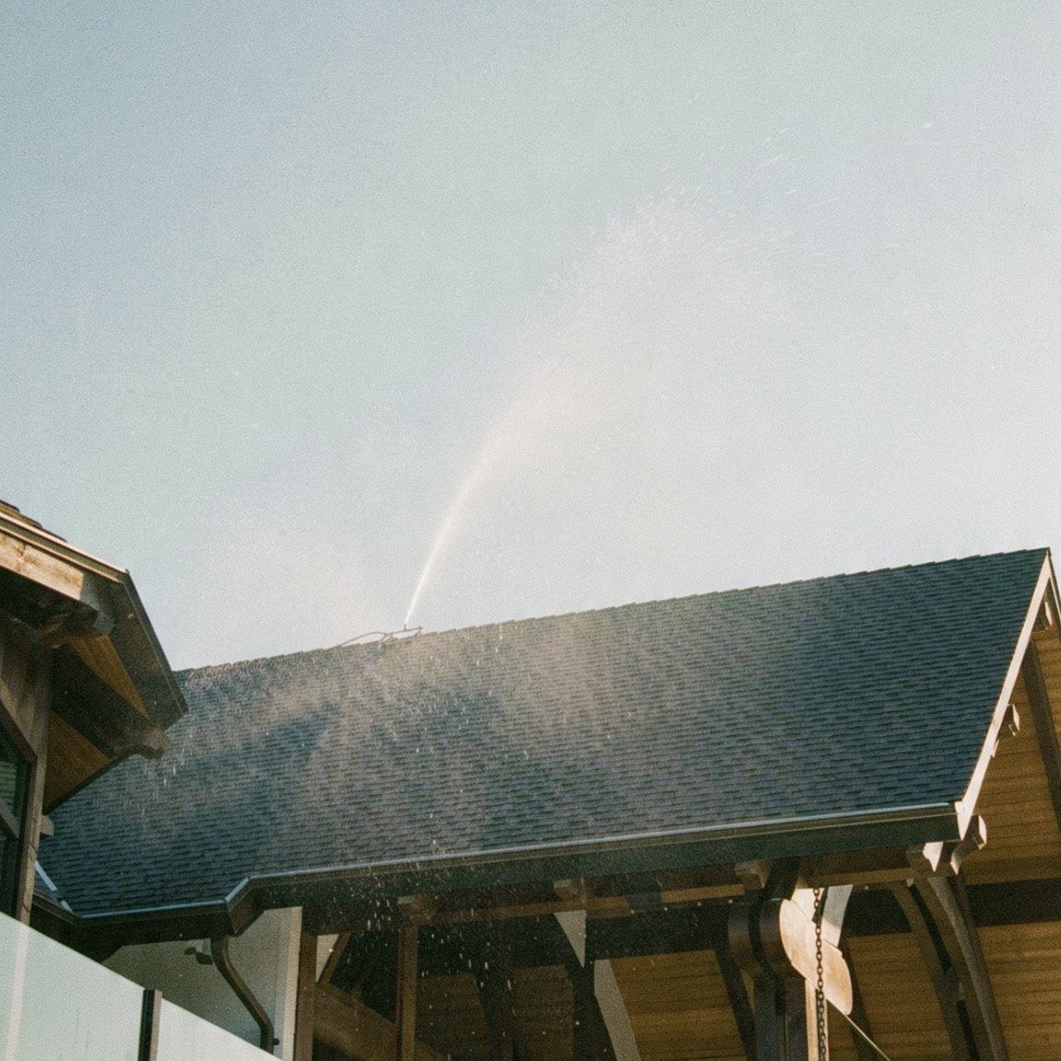 Rooftop wildfire sprinkler spraying a tall water arc over a shingled Okanagan home roof
